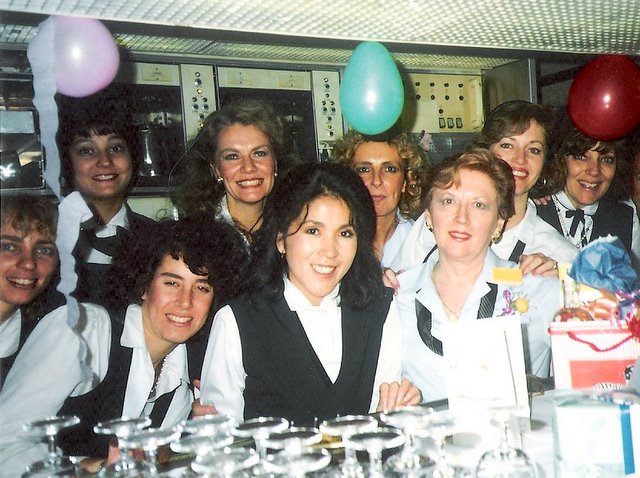 1980s Purser, Peggy Catalano,(wearing a yellow rose) on her last working flight before retirement poses in the Economy Class galley of a Pan Am 747 with fellow crew members.  Judy Skartvedt is standing behind Peggy with hands on Peggy's shoulders. 